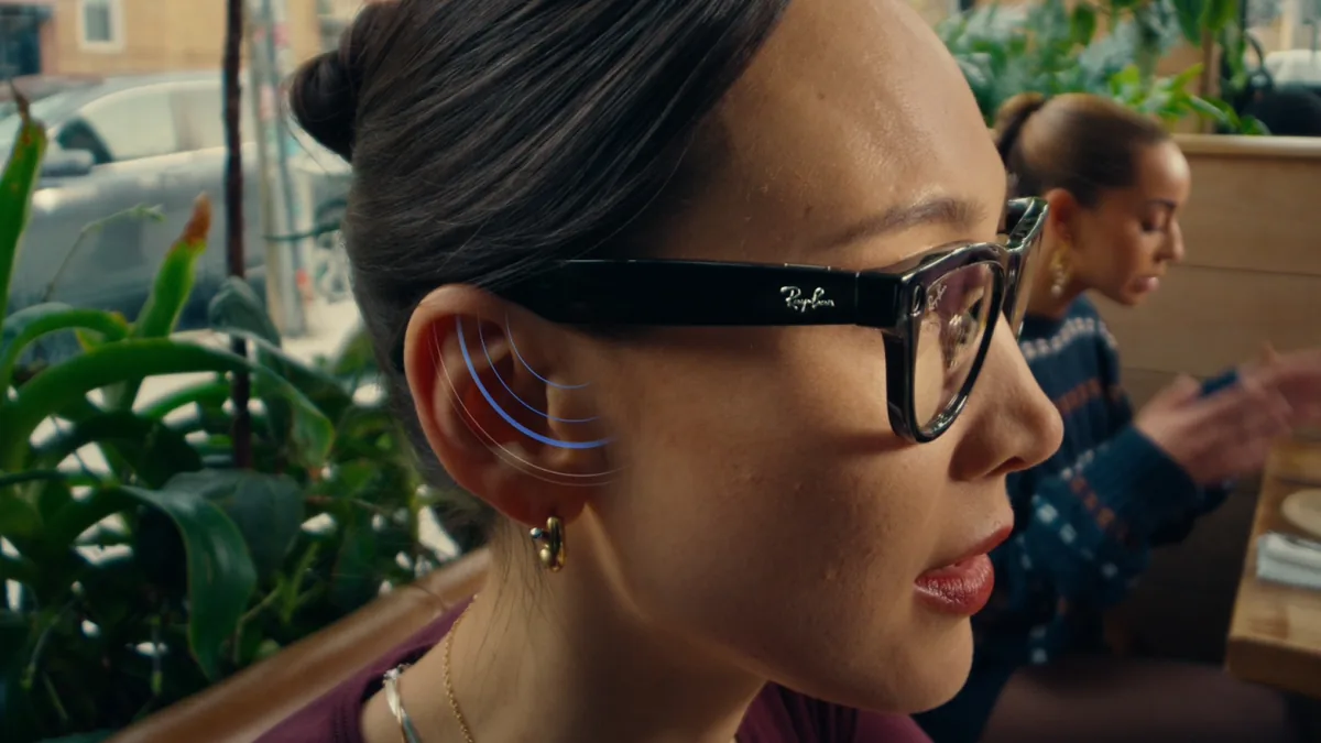 A woman wearing Ray-Ban Meta glasses faces another person during a conversation in a busy setting.